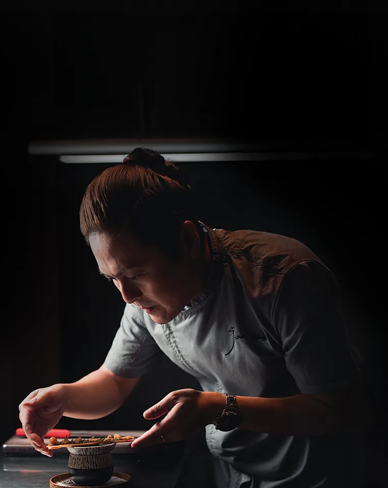 Chef Tyrell Joon plating a dish under a single low light in the Café Café kitchen
