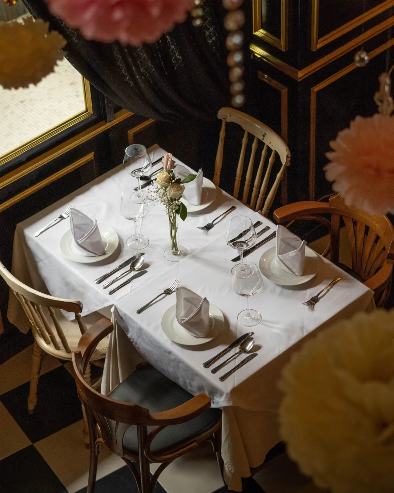 A table for two laid with white linen, silver and a single rose in a bud vase, seen from above against the restaurant's black and white checkerboard floor
