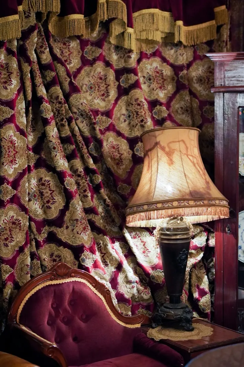 A fringed table lamp beside a tufted velvet chair, against a heavy burgundy and gold damask curtain
